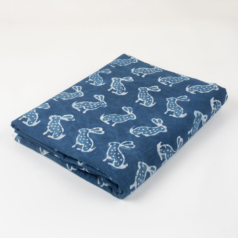 May include: Folded fabric with a navy blue background and a repeating pattern of white rabbits. The rabbits are stylized with dotted bodies and long ears. The fabric is folded neatly, showing the pattern on the top layer.