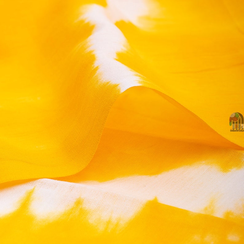 May include: Close-up of a yellow fabric with white tie-dye patterns. The fabric is soft and flowing, creating a gentle wave.