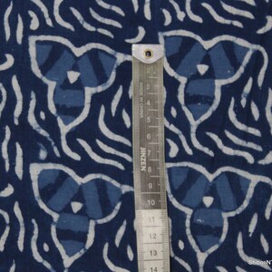 May include: Indigo blue cotton fabric with a white and blue patterned design. The fabric has a repeating pattern of a stylized flower with a white outline. A ruler is included for scale.