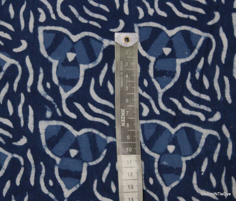 May include: Indigo blue cotton fabric with a white and blue patterned design. The fabric has a repeating pattern of a stylized flower with a white outline. A ruler is included for scale.