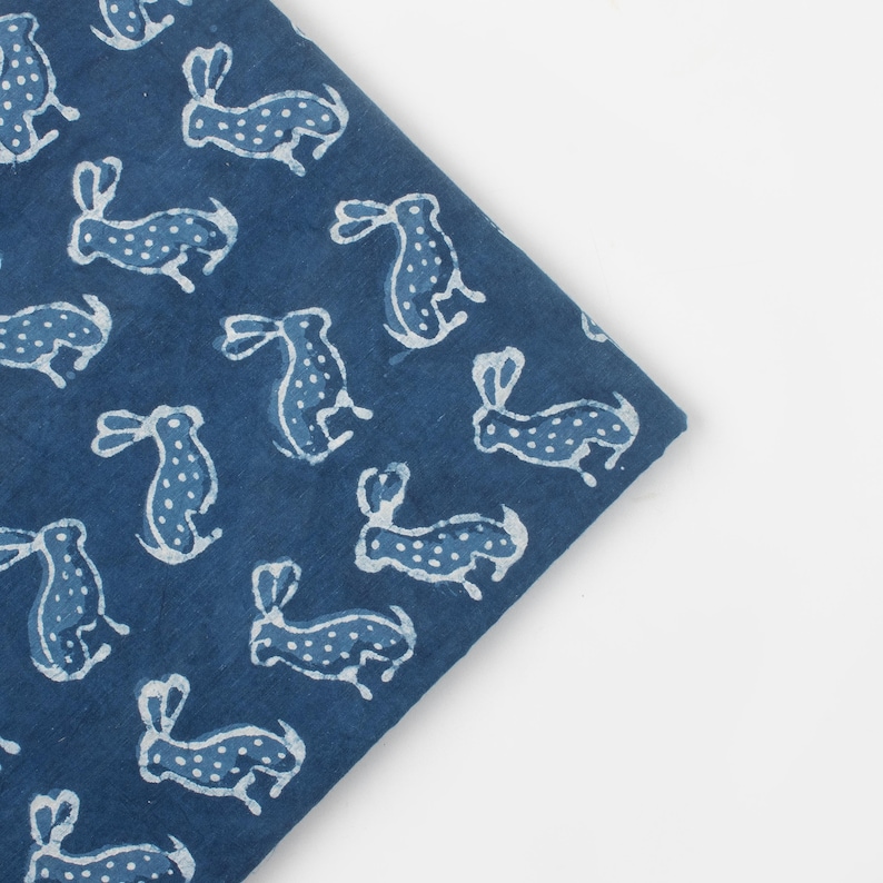 May include: A folded piece of indigo blue fabric with a repeating pattern of white dotted rabbits. The rabbits are in various poses, creating a whimsical and playful design. The fabric is likely cotton or linen.