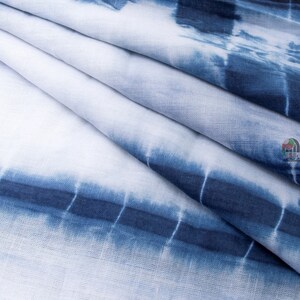 Op de afbeelding: Close-up van een witte stof met een blauw tie-dye patroon. De stof is in meerdere lagen gevouwen, waardoor een textuur ontstaat.