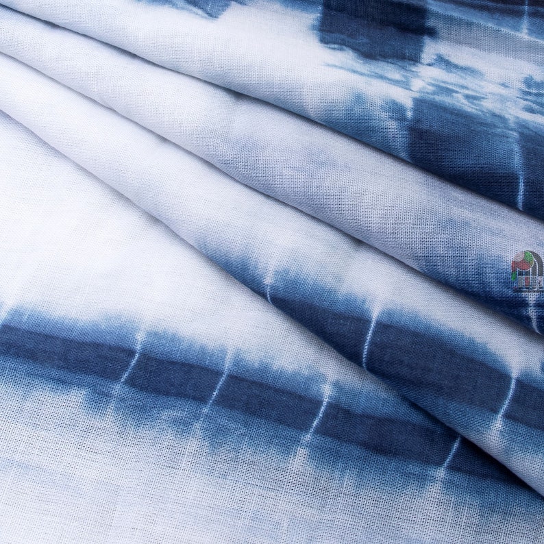 Op de afbeelding: Close-up van een witte stof met een blauw tie-dye patroon. De stof is in meerdere lagen gevouwen, waardoor een textuur ontstaat.