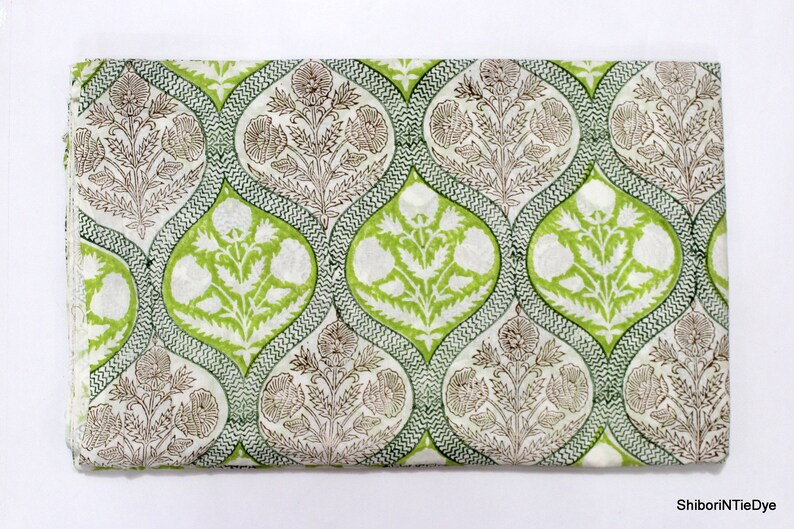May include: White cotton fabric with a green and brown floral print. The pattern features repeating teardrop shapes with a floral design inside. The fabric is folded in half.