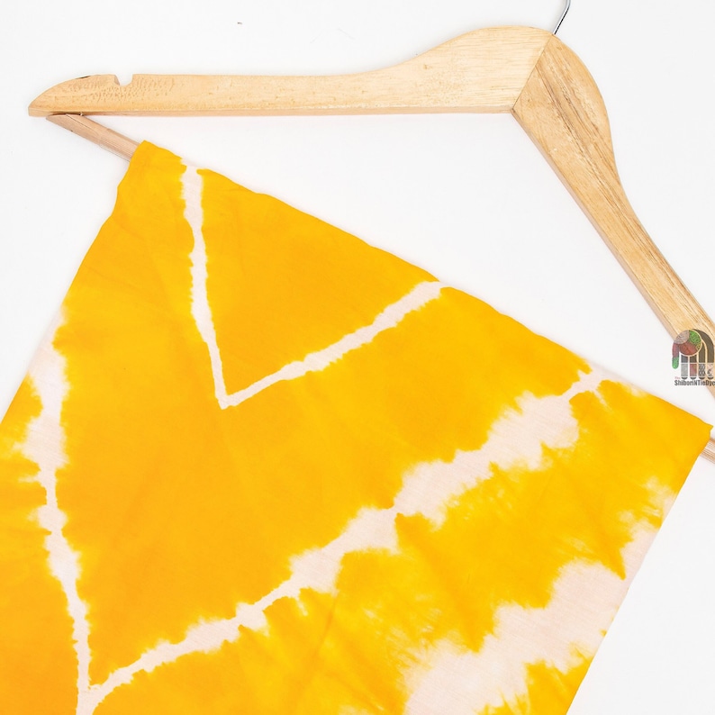 May include: A yellow and white tie-dye fabric hanging on a wooden hanger. The fabric has a swirling pattern of yellow and white.