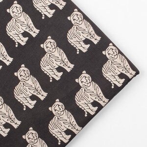 May include: Black fabric with a repeating pattern of cream-colored tiger illustrations. The tigers are stylized with detailed stripes and a seated pose. The fabric is folded, showing the pattern's edge.