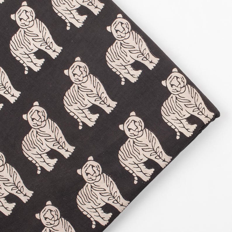 May include: Black fabric with a repeating pattern of cream-colored tiger illustrations. The tigers are stylized with detailed stripes and a seated pose. The fabric is folded, showing the pattern's edge.