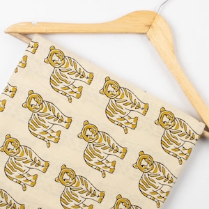May include: Cream-colored fabric with a repeating pattern of stylized yellow tigers. The tigers are depicted in a relaxed pose with striped bodies. The fabric is draped over a wooden hanger, suggesting it's ready for use in sewing or crafting projects.