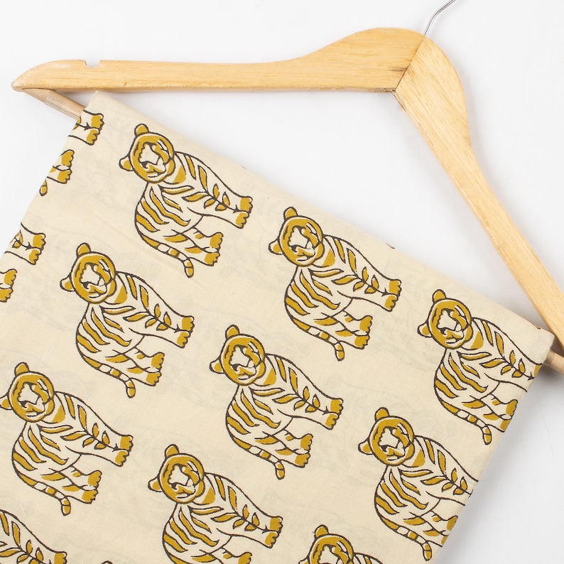 May include: Cream-colored fabric with a repeating pattern of stylized yellow tigers. The tigers are depicted in a relaxed pose with striped bodies. The fabric is draped over a wooden hanger, suggesting it's ready for use in sewing or crafting projects.