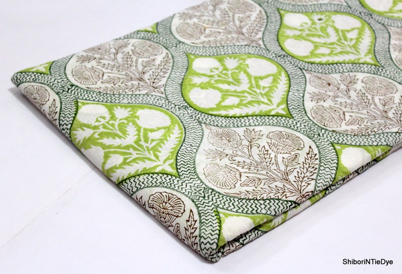 May include: White cotton fabric with a green and brown floral print. The print features a repeating pattern of teardrop shapes with floral designs inside. The fabric is folded in half.