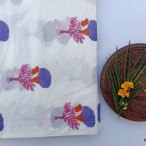 Puede incluir: Tela blanca con un patr&oacute;n repetido de dise&ntilde;os florales estilizados en morado, naranja y rosa. Un plato de madera con detalles tallados contiene vegetaci&oacute;n y flores amarillas. La tela est&aacute; doblada.