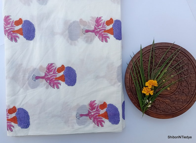 Puede incluir: Tela blanca con un patr&oacute;n repetido de dise&ntilde;os florales estilizados en morado, naranja y rosa. Un plato de madera con detalles tallados contiene vegetaci&oacute;n y flores amarillas. La tela est&aacute; doblada.