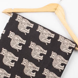 May include: Black fabric with a repeating pattern of beige tiger illustrations. The fabric is draped over a wooden hanger. The tigers are stylized with detailed outlines and stripes, creating a bold and eye-catching design. Ideal for apparel or home decor projects.