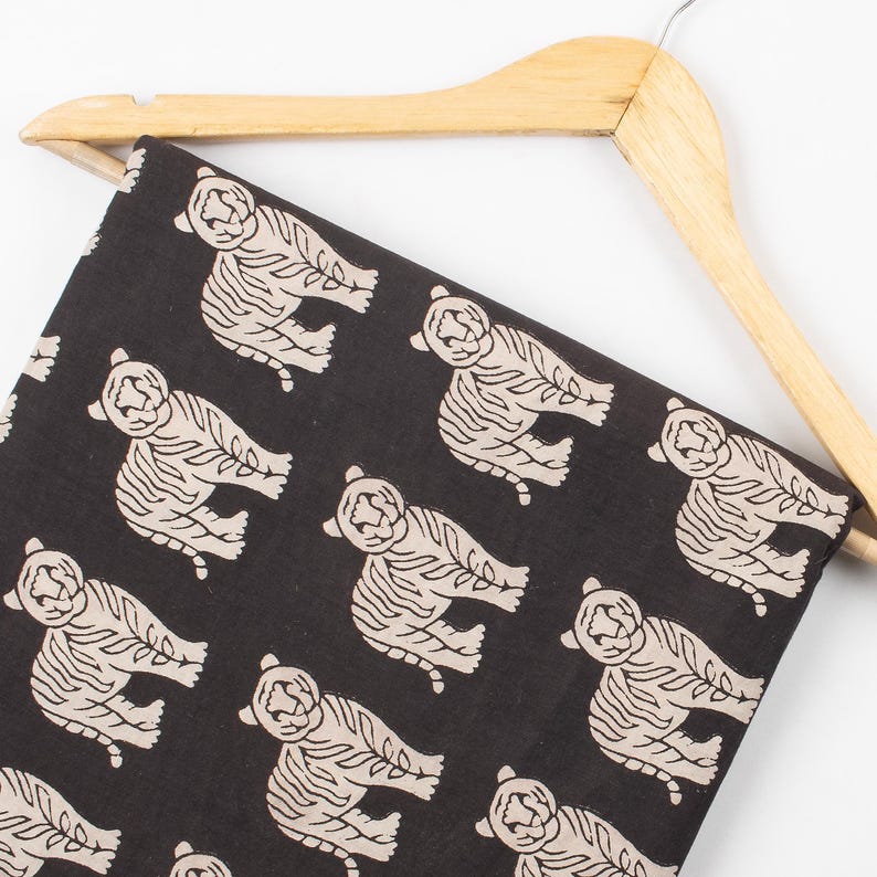 May include: Black fabric with a repeating pattern of beige tiger illustrations. The fabric is draped over a wooden hanger. The tigers are stylized with detailed outlines and stripes, creating a bold and eye-catching design. Ideal for apparel or home decor projects.
