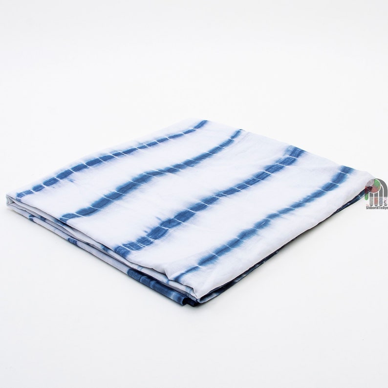 Op de afbeelding: Een witte stof met blauwe tie-dye strepen gevouwen in de helft. De stof heeft een zachte, vloeiende textuur.