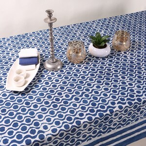 May include: A white tablecloth with a blue wave pattern. The tablecloth is set with a silver candlestick, two clear glass candle holders, a small potted plant, and a white rectangular serving tray with two small white bowls.