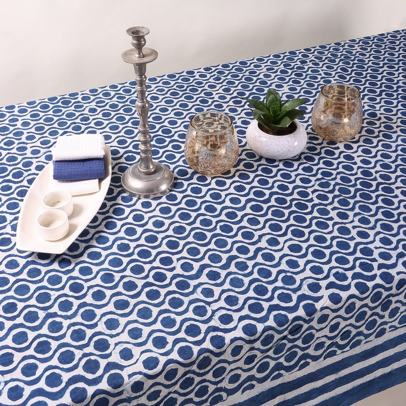 May include: A white tablecloth with a blue wave pattern. The tablecloth is set with a silver candlestick, two clear glass candle holders, a small potted plant, and a white rectangular serving tray with two small white bowls.