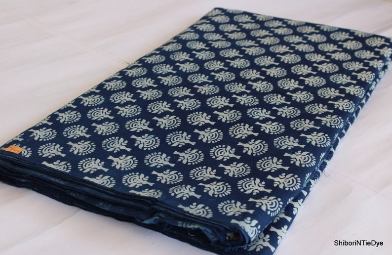 May include: A folded piece of indigo blue cotton fabric with a white floral pattern. The fabric is folded in thirds and has a white tag with the text "ShiboriNTieDye" on the bottom right corner.