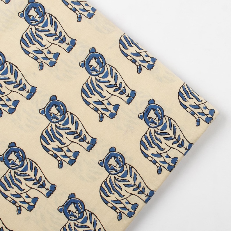 May include: Cream-colored fabric with a repeating pattern of blue tiger illustrations. The tigers are stylized with striped bodies and detailed features. The fabric is folded, showing the pattern's continuity.
