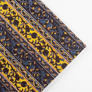 May include: Close-up of a fabric with a repeating pattern of floral designs. The design features alternating stripes of yellow, navy blue, and black. The floral elements are in navy blue and black, with white and red accents. The fabric is folded at a diagonal angle.