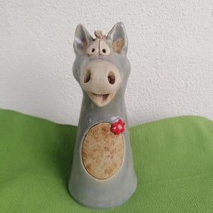 Könnte beinhalten: Eine skurrile Esel-Figur aus Keramik in Grau-, Beige- und Rottönen. Der Esel hat ein lächelndes Gesicht, große Ohren und eine rote Blume. Die Figur hat einen abgerundeten Körper mit einem strukturierten, ovalen Fleck.