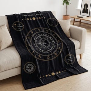 May include: A black throw blanket with a celestial map design in gold and white. The blanket features zodiac symbols, phases of the moon, and the text "THE CELESTIAL MAP OF". The blanket is draped over a beige sofa.