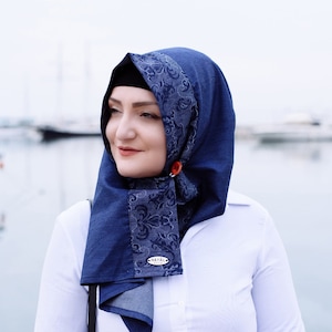 May include: A woman wearing a white button-down shirt with rolled up sleeves and a blue denim hijab with a floral pattern. The hijab has a silver tag that says "HAYAL" on it. She is wearing a ring on her finger.