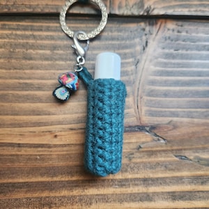 Pode incluir: Um porta-chaves de crochê turquesa com um tubo de bálsamo labial branco dentro. O porta-chaves tem um anel prateado e um amuleto de cogumelo colorido.