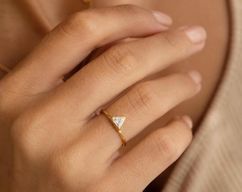 Op de afbeelding: Een gouden ring met een enkele, kleine, heldere, driehoekig geslepen edelsteen in een zetting. De ringband is dun en gepolijst, met een eenvoudig ontwerp dat de geometrische vorm van de steen in het midden van de vinger benadrukt.