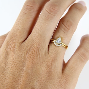 Könnte beinhalten: Eine Hand, die zwei goldene Ringe trägt. Der Hauptring hat einen birnenförmigen, klaren Edelstein. Ein zweiter Ring, ebenfalls aus Gold, ist ein gebogenes Band mit kleinen, klaren Edelsteinen. Die Ringe sind vor einem weißen Hintergrund platziert.