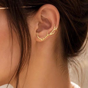 Peut inclure: Un ear cuff doré en forme de bois de cerf ramifié. Ce bijou épouse la courbe naturelle du pavillon de l'oreille, présentant une finition métallique lisse qui imite la structure organique et bifurquée d'un bois de cerf en une seule pièce.