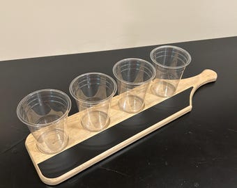 Könnte beinhalten: Ein helles Holz-Servierbrett mit einer langen, schwarzen, rechteckigen Einlage im Tafel-Stil in der Mitte. Vier durchsichtige Einweg-Plastikbecher sind in kreisförmigen Aussparungen entlang der Länge des Holzbretts angeordnet.