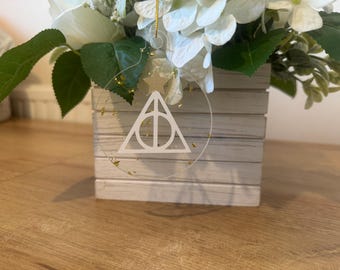 May include: A clear, circular ornament with a white Deathly Hallows symbol, gold glitter, and a gold star. The ornament hangs in front of a white floral arrangement in a wooden box.