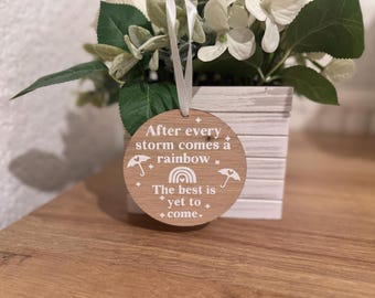 May include: Wooden round sign with the text "After every storm comes a rainbow. The best is yet to come."  The sign has a white background and white text with a rainbow and two umbrellas.