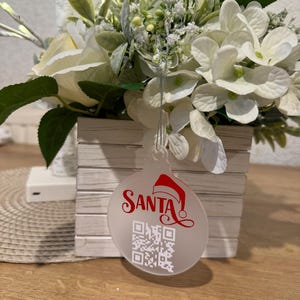 May include: A clear, round Christmas bauble with the word "SANTA" in red script and a Santa hat graphic. A QR code is printed below the text. The bauble hangs in front of a white floral arrangement.
