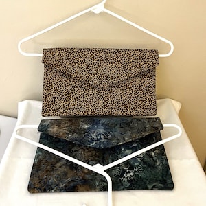 May include: Two fabric clutch purses. The top purse has a tan and brown leopard print pattern. The bottom purse has a blue, green, and brown floral pattern. Both purses are displayed on white hangers.
