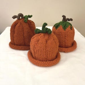 May include: Three hand-knitted pumpkin-shaped hats in orange yarn. Each hat features a rolled brim, a textured ribbed body, and a green crocheted stem with a small curly vine detail on top. The hats are arranged in a triangular formation.