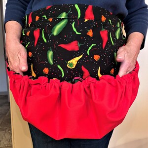 May include: A black apron with a vibrant chili pepper pattern, featuring red, green, and yellow peppers against a speckled background. The apron has a bright red gathered pocket at the bottom, offering a bold contrast.
