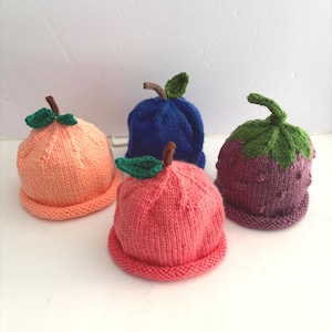 May include: Four knitted baby hats in the shape of fruits. The hats are in shades of peach, pink, blue, and purple, with green leaf and brown stem details. The hats are designed to resemble an orange, apple, blueberry, and strawberry.