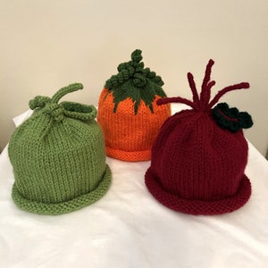 May include: Three knitted hats in the shape of vegetables. One is green, resembling a pea pod. The second is orange with green leaves, like a pumpkin. The third is burgundy, with a green leaf and red tendrils, resembling a beet.