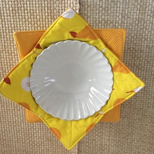 May include: A white, scalloped-edge plate rests on a yellow and orange quilted pot holder. The pot holder has a star shape with a pear pattern on the top layer and a polka dot pattern on the bottom layer. The background is a neutral woven fabric.