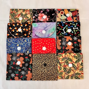 May include: A collection of small, fabric pouches with various patterns. Designs include holly, floral, candy corn, bone, gingerbread, and leopard print. Each pouch has a curved flap and a white or black snap closure. The pouches are arranged on a white surface.