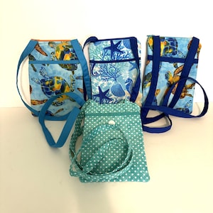 May include: Four rectangular fabric crossbody bags with long straps. Three bags feature blue-toned sea life prints including turtles, starfish, and coral. One bag is teal with a white polka dot pattern and a single white snap closure on the front.