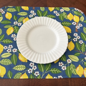 May include: A rectangular placemat with a blue background featuring a lemon and floral pattern. A white, round plate with a ridged edge sits on top. The lemons are yellow, and the leaves are green.