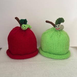 May include: Two knitted baby hats, one red and one green, designed to resemble apples. Each hat features a brown stem, green leaf, and a smiling caterpillar. The hats are made of a chunky knit material.