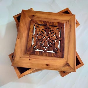 Peut inclure: Une boîte en bois avec un motif sculpté sur le couvercle. La boîte est en bois brun clair et a une forme carrée. La sculpture est complexe et détaillée, et elle présente un motif floral.