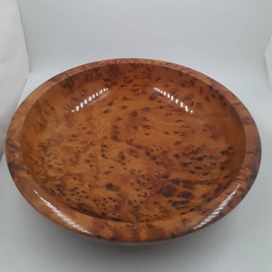May include: A circular wooden bowl with a polished surface. The bowl is crafted from light brown wood, showcasing a distinctive, swirling grain. The interior is smooth and reflective, and the rim is rounded.