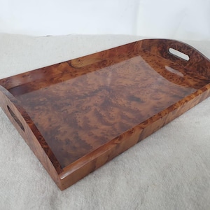 Peut inclure: Plateau de service rectangulaire en bois avec une finition brillante. Le plateau présente un motif de loupe de bois brun chaud et des poignées découpées. Une pièce décorative pour servir des aliments ou exposer des objets.