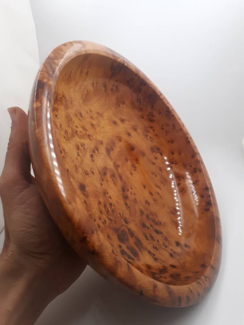 May include: An oval wooden bowl with a polished finish. The bowl is made of light brown wood with dark brown knots and swirls. The interior is smooth and glossy. The bowl is held in a hand.
