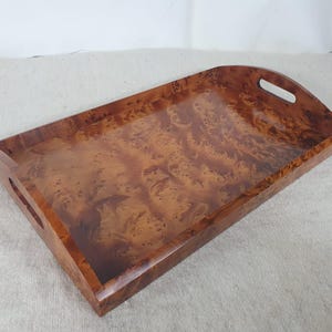 Peut inclure: Un plateau de service rectangulaire en bois avec un motif de grain de loupe brun chaud et riche. Le plateau a des poignées incurvées à chaque extrémité et est conçu pour transporter des articles. Le plateau mesure environ 46 cm de long et 30 cm de large.